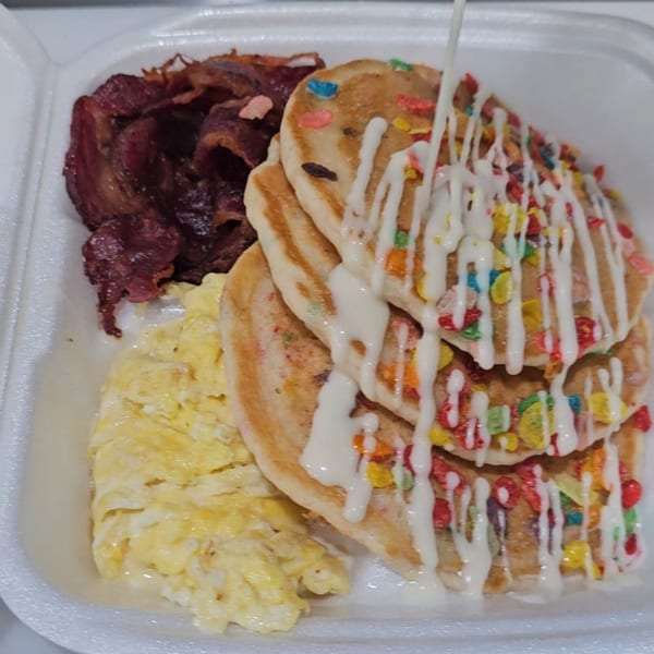 Brunch Bro's pancakes with colorful cereal, white icing, scrambled eggs, and bacon in a takeout container.