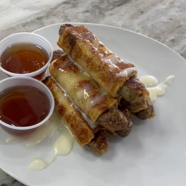 French toast wrapped sausages with glaze and two cups of syrup on a white plate.