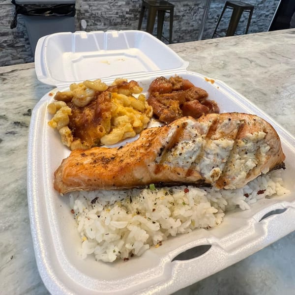 Soul food takeout with salmon, rice, mac and cheese, and candied yams in a styrofoam container.