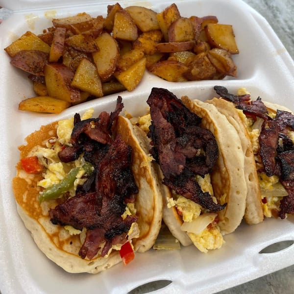 Soul food breakfast with pancakes, scrambled eggs, bacon, and seasoned home fries in a takeout container.