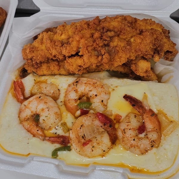 Fried catfish and shrimp and grits with peppers and spices in a white takeout container.
